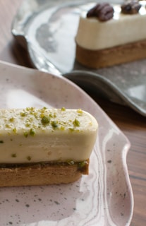 A rectangular dessert with a cream-colored mousse topped with chopped green pistachios, set on a biscuit base served on a speckled ceramic plate. In the background, another similar dessert garnished with pecans sits on a dark plate.