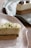 A rectangular dessert with a cream-colored mousse topped with chopped green pistachios, set on a biscuit base served on a speckled ceramic plate. In the background, another similar dessert garnished with pecans sits on a dark plate.