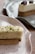 A rectangular dessert with a cream-colored mousse topped with chopped green pistachios, set on a biscuit base served on a speckled ceramic plate. In the background, another similar dessert garnished with pecans sits on a dark plate.