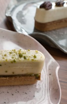 A rectangular dessert with a cream-colored mousse topped with chopped green pistachios, set on a biscuit base served on a speckled ceramic plate. In the background, another similar dessert garnished with pecans sits on a dark plate.