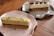 Two rectangular pastries with a creamy, white topping are placed on different plates on a wooden surface. One is garnished with chopped nuts and placed on a pink plate, while the other is topped with whole nuts and served on a grey plate.