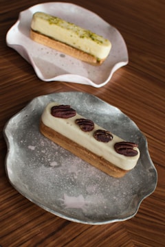 Two elegant rectangular pastries are displayed on artistic plates. The front pastry is topped with pecans and rests on a rustic silver plate. Behind it, another pastry garnished with pistachios is placed on a pink and white ceramic plate. Both pastries are set on a wooden surface that adds warmth to the composition.