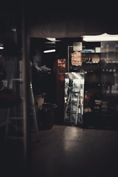 A dimly lit interior with a display window featuring multiple shoes arranged on shelves. A metal ladder is visible in the center, reflecting the light from outside. The scene includes a mix of dark shadows and soft lighting. A 'No Smoking' sign and decorative elements are present.