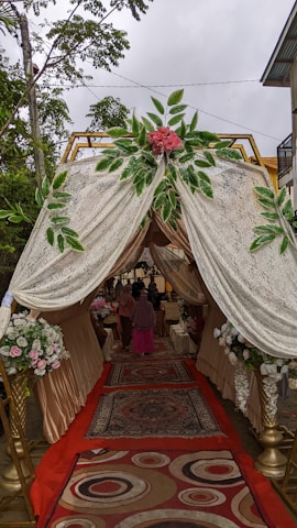 A canopy with ornate drapes and floral arrangements at the entrance. The interior features carpeted flooring with intricate patterns, leading to an event space where people are gathered. The canopy is adorned with lush greenery and pink flowers, creating an elegant and welcoming atmosphere.
