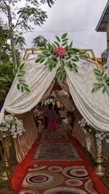 A canopy with ornate drapes and floral arrangements at the entrance. The interior features carpeted flooring with intricate patterns, leading to an event space where people are gathered. The canopy is adorned with lush greenery and pink flowers, creating an elegant and welcoming atmosphere.