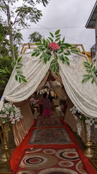 A canopy with ornate drapes and floral arrangements at the entrance. The interior features carpeted flooring with intricate patterns, leading to an event space where people are gathered. The canopy is adorned with lush greenery and pink flowers, creating an elegant and welcoming atmosphere.