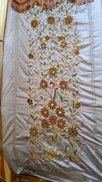 A detailed embroidery pattern on a fabric showcasing floral designs. The pattern features large, symmetrical flowers with petals in shades of orange, complemented by green leaves and stems. The fabric has a shiny appearance with a decorative border along the top edge, adorned with tassels.