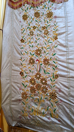 A detailed embroidery pattern on a fabric showcasing floral designs. The pattern features large, symmetrical flowers with petals in shades of orange, complemented by green leaves and stems. The fabric has a shiny appearance with a decorative border along the top edge, adorned with tassels.