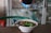 A bowl of noodle soup is being prepared with a dark sauce poured from a bottle. The dish contains green vegetables and sliced ingredients, and a spoon and fork rest inside the bowl. In the background, some shelves with items are visible.