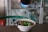 A bowl of noodle soup is being prepared with a dark sauce poured from a bottle. The dish contains green vegetables and sliced ingredients, and a spoon and fork rest inside the bowl. In the background, some shelves with items are visible.