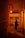 A person stands alone in a narrow alleyway illuminated by a warm orange glow. They are holding a cup and wearing a hat, standing next to a building with visible electrical wiring and an air conditioning unit. The scene conveys a sense of solitude and contemplation.