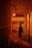 A person stands alone in a narrow alleyway illuminated by a warm orange glow. They are holding a cup and wearing a hat, standing next to a building with visible electrical wiring and an air conditioning unit. The scene conveys a sense of solitude and contemplation.