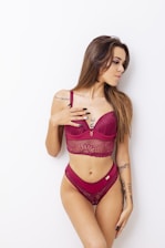 A woman poses against a white background wearing a burgundy lace bralette and matching bottoms. Her long hair is styled naturally, and she has several tattoos on her arms. She gazes to the side with a relaxed demeanor.