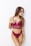 A woman poses against a white background wearing a burgundy lace bralette and matching bottoms. Her long hair is styled naturally, and she has several tattoos on her arms. She gazes to the side with a relaxed demeanor.