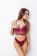 A woman poses against a white background wearing a burgundy lace bralette and matching bottoms. Her long hair is styled naturally, and she has several tattoos on her arms. She gazes to the side with a relaxed demeanor.