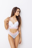 A woman with long brown hair stands against a plain white background. She is wearing light blue and pink lace lingerie. There are visible tattoos on her arms and body, and she has manicured black nails.