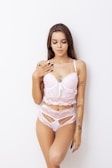 A woman with long hair poses against a plain white background wearing a pale pink lace lingerie set with transparent accents. Her body is adorned with several tattoos, and she has her fingers gently touching her collarbone.