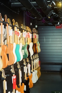 A row of electric guitars is displayed hanging on a dark wall, each guitar boasting different colors such as turquoise, natural wood, black, white, and orange. The setting appears to be a music store or showroom with industrial overhead pipes and spotlights.