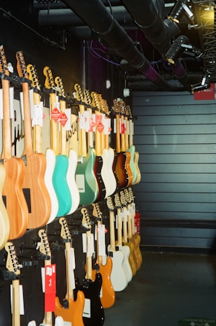A row of electric guitars is displayed hanging on a dark wall, each guitar boasting different colors such as turquoise, natural wood, black, white, and orange. The setting appears to be a music store or showroom with industrial overhead pipes and spotlights.