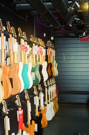 A row of electric guitars is displayed hanging on a dark wall, each guitar boasting different colors such as turquoise, natural wood, black, white, and orange. The setting appears to be a music store or showroom with industrial overhead pipes and spotlights.