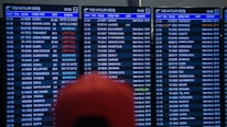 Three airport departure boards displaying various international flight details such as times, destinations, gate numbers, and status updates. The information is shown in white text on a dark background, with special indications like 'Last Call' and 'Boarding' highlighted in red. The text is in both English and another language. A blurry red object is partially visible in front of the boards.