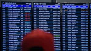 Three airport departure boards displaying various international flight details such as times, destinations, gate numbers, and status updates. The information is shown in white text on a dark background, with special indications like 'Last Call' and 'Boarding' highlighted in red. The text is in both English and another language. A blurry red object is partially visible in front of the boards.
