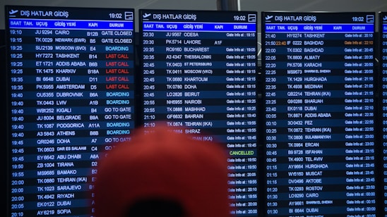 Three airport departure boards displaying various international flight details such as times, destinations, gate numbers, and status updates. The information is shown in white text on a dark background, with special indications like 'Last Call' and 'Boarding' highlighted in red. The text is in both English and another language. A blurry red object is partially visible in front of the boards.