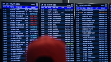 Three airport departure boards displaying various international flight details such as times, destinations, gate numbers, and status updates. The information is shown in white text on a dark background, with special indications like 'Last Call' and 'Boarding' highlighted in red. The text is in both English and another language. A blurry red object is partially visible in front of the boards.