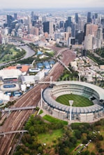 An aerial view of a large cricket stadium with a circular design, surrounded by train tracks and a river winding through the skyline of a bustling urban city. Skyscrapers line the horizon, and there are green parks and trees scattered throughout the landscape, contributing to the city's blend of nature and urban architecture.