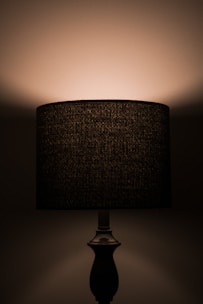 A dimly lit lamp with a dark, textured lampshade casting soft light onto the wall behind it. The base is simple and elegant, enhancing the overall subtlety of the setup.
