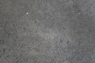 A textured concrete surface with a slightly rough appearance and scattered small stones. The surface contains a few cracks and a spot of different color near the center.
