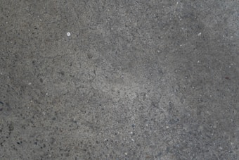A textured concrete surface with a slightly rough appearance and scattered small stones. The surface contains a few cracks and a spot of different color near the center.