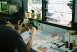 A person is sitting at a desk engaged in a creative activity. The desk is cluttered with various art supplies such as paints, brushes, and containers. There is a window in the background, allowing natural light to fill the space. Several plants and a computer monitor are visible, contributing to a casual and artistic atmosphere.