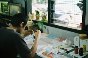 A person is sitting at a desk engaged in a creative activity. The desk is cluttered with various art supplies such as paints, brushes, and containers. There is a window in the background, allowing natural light to fill the space. Several plants and a computer monitor are visible, contributing to a casual and artistic atmosphere.