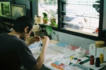A person is sitting at a desk engaged in a creative activity. The desk is cluttered with various art supplies such as paints, brushes, and containers. There is a window in the background, allowing natural light to fill the space. Several plants and a computer monitor are visible, contributing to a casual and artistic atmosphere.