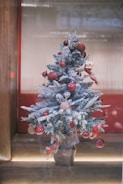 A small artificial Christmas tree decorated with red ornaments, ribbons, and snow-like flocking is placed inside a windowed display or alcove. The tree stands on a wooden floor and has a rustic, burlap-covered base. The background features frosted glass and red paneling, adding a festive ambiance.
