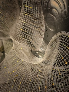 A close-up view of a white mesh fabric, intricately layered and forming wavy, flowing patterns against a background that appears metallic or reflective.