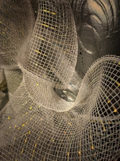 A close-up view of a white mesh fabric, intricately layered and forming wavy, flowing patterns against a background that appears metallic or reflective.