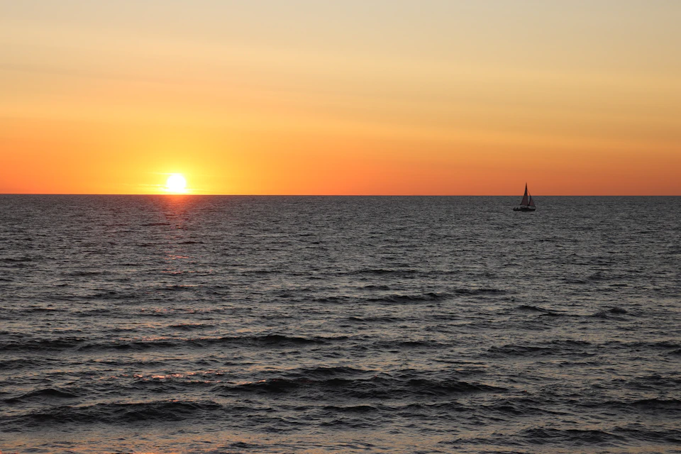 Sunset over ocean with sailboat in the distance, casting warm glow across the sky