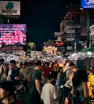 A bustling night market scene with a large crowd of people walking and shopping along a narrow street lined with bright, colorful signs and vendor stalls. Various signs in different languages illuminate the area, reflecting the lively atmosphere. A mix of locals and tourists can be seen browsing the stalls, engaged in conversation, or looking at their phones.