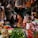 A bustling market stall filled with a variety of goods including vegetables like cabbages, limes, and green onions. Packaged items hang above, and there is a mix of spices and food products in bottles and bags. A person is seated, tending to the stall, looking contemplative.