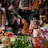 A bustling market stall filled with a variety of goods including vegetables like cabbages, limes, and green onions. Packaged items hang above, and there is a mix of spices and food products in bottles and bags. A person is seated, tending to the stall, looking contemplative.
