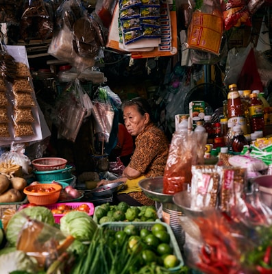 A bustling market stall filled with a variety of goods including vegetables like cabbages, limes, and green onions. Packaged items hang above, and there is a mix of spices and food products in bottles and bags. A person is seated, tending to the stall, looking contemplative.