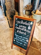 A wooden-framed chalkboard placed on a stone floor with text in both Thai and English, instructing customers to order drinks or food before entering the cafe's areas. In the background, there's a stone sculpture and a potted plant with dried flowers.