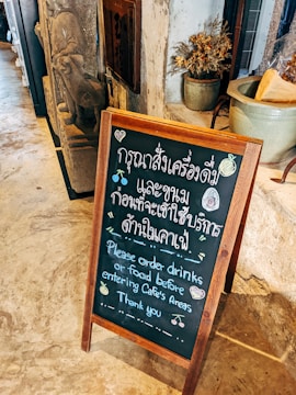 A wooden-framed chalkboard placed on a stone floor with text in both Thai and English, instructing customers to order drinks or food before entering the cafe's areas. In the background, there's a stone sculpture and a potted plant with dried flowers.