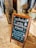 A wooden-framed chalkboard placed on a stone floor with text in both Thai and English, instructing customers to order drinks or food before entering the cafe's areas. In the background, there's a stone sculpture and a potted plant with dried flowers.