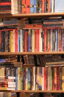 A variety of books is arranged on several wooden shelves, featuring multiple colors and genres. The books are stacked both vertically and horizontally, with some titles clearly visible, including classic and contemporary works.