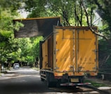 A large yellow truck is parked on a tree-lined suburban street. Trees arch overhead, with their branches reaching toward the truck. The truck's rear doors are prominently visible, and there appears to be signage with contact information on them. In the distance, a small white car is parked on the side of the road.