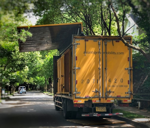 A large yellow truck is parked on a tree-lined suburban street. Trees arch overhead, with their branches reaching toward the truck. The truck's rear doors are prominently visible, and there appears to be signage with contact information on them. In the distance, a small white car is parked on the side of the road.