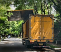 A large yellow truck is parked on a tree-lined suburban street. Trees arch overhead, with their branches reaching toward the truck. The truck's rear doors are prominently visible, and there appears to be signage with contact information on them. In the distance, a small white car is parked on the side of the road.